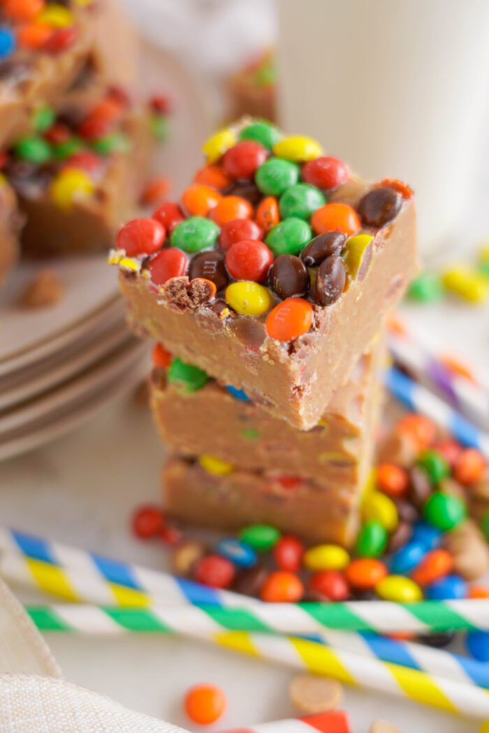 Stack of peanut butter fudge topped with colorful M&Ms chocolates, with more scattered around on a white plate.