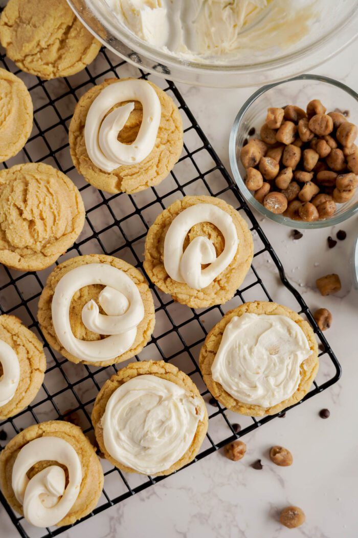 A cooling rack with frosted sugar cookies, some featuring swirled frosting and others spread evenly. Nearby are bowls brimming with frosting and chocolate-covered nuts on a white surface. Try our Easy Cookie Dough Cookies Recipe to recreate this delightful scene at home.