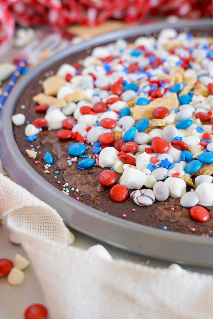 A close-up view of a chocolate brownie pizza topped with red, white, and blue candies, marshmallows, and sprinkles.