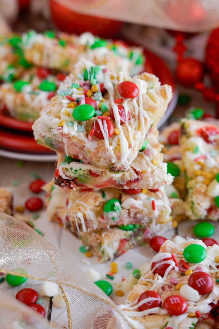 A stack of Christmas Cookie bars topped with white chocolate drizzle, colorful candies, and sprinkles, resembling a festive M&M Christmas Cookie Bars Recipe, with more decorated bars and festive decorations in the background.