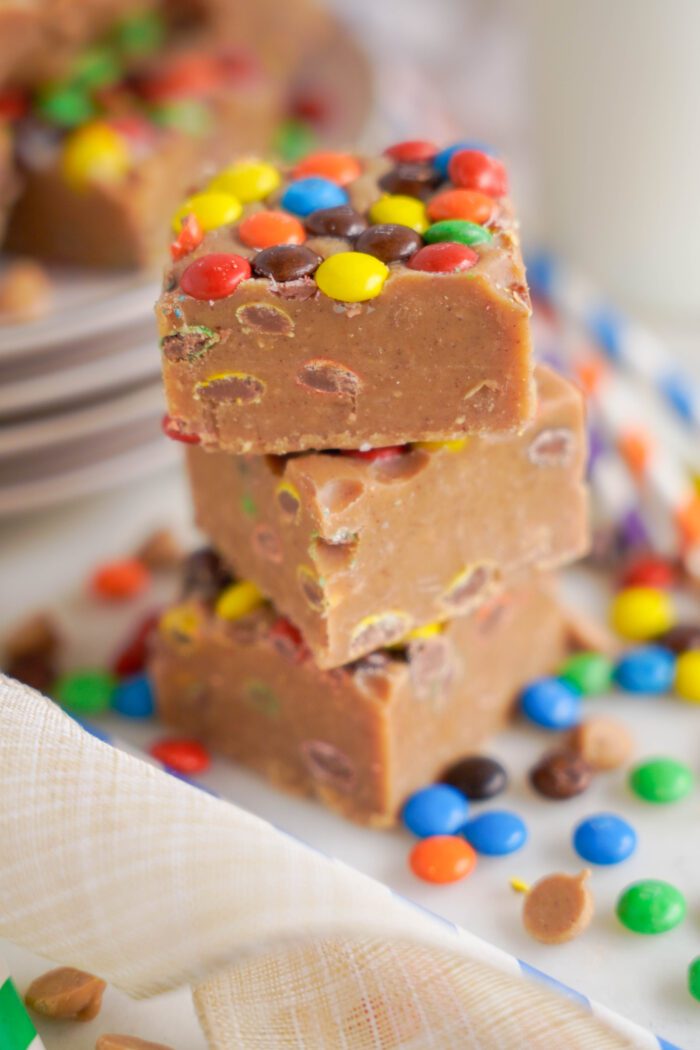 Stack of fudge pieces with colorful M&M pieces mixed in, displayed on a white plate with more M&Ms scattered around.
