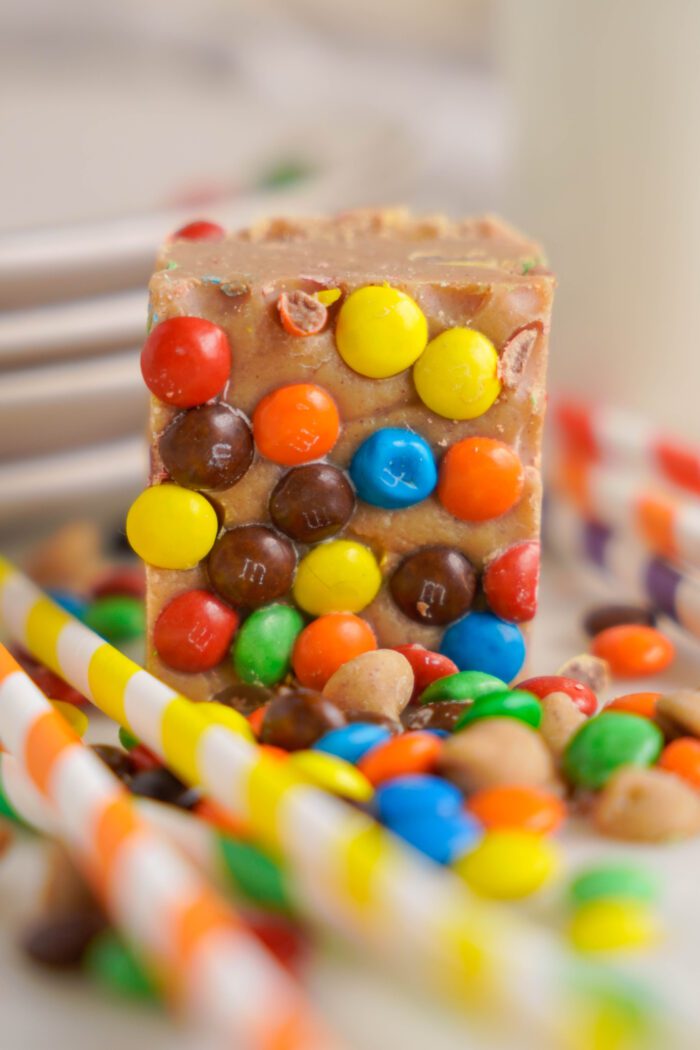 A close-up of a Peanut Butter Fudge square surrounded by colorful M&Ms and striped straws.
