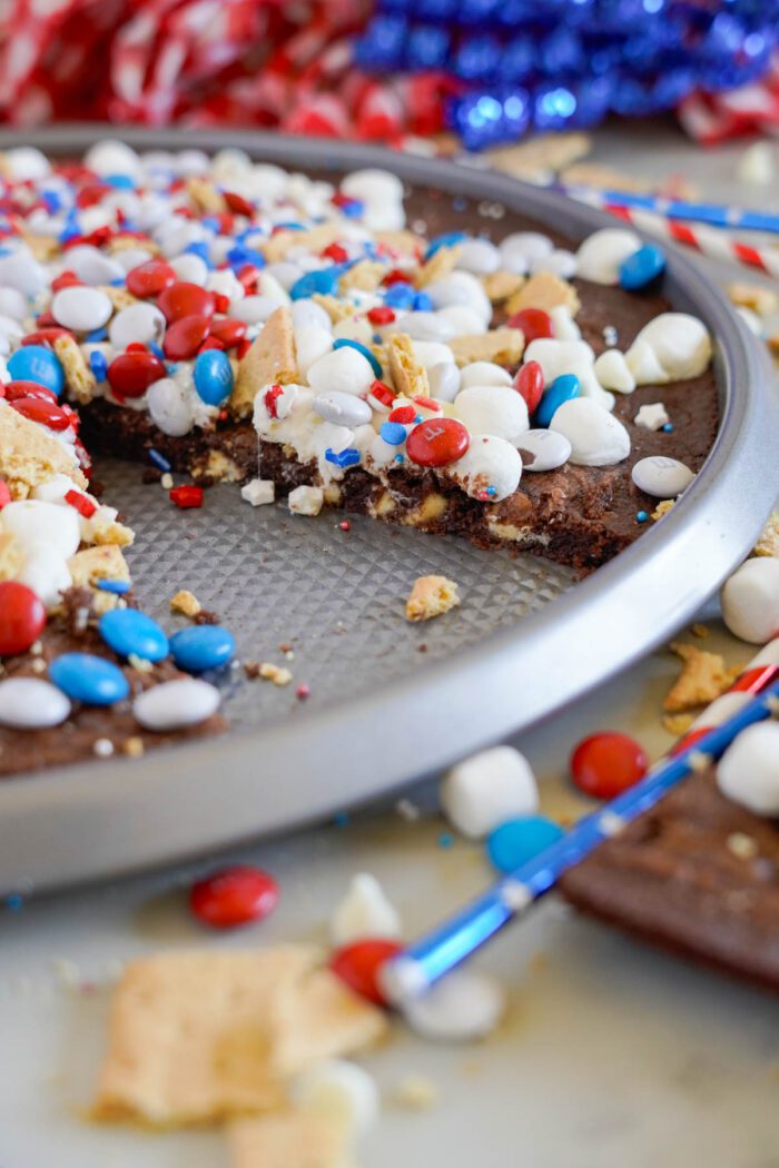 A close-up of a partially eaten brownie pizza topped with red, white, and blue candies and marshmallows on a round baking sheet.