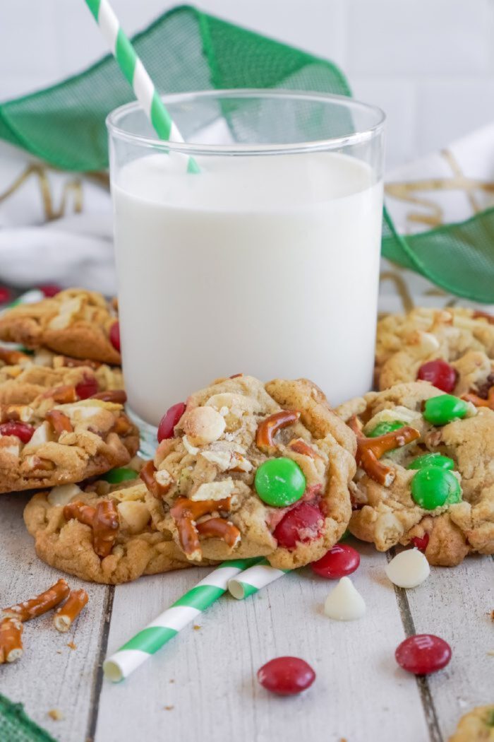 A glass of milk with a striped straw is surrounded by cookies containing red, green, and white candy pieces, pretzels, and white chocolate chips; a green and white striped straw lies on the surface.