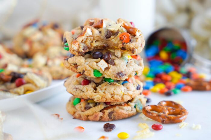 A stack of three cookies with colorful candy pieces and chocolate chips. Nearby, scattered candies and a pretzel are visible. A blurred plate with more cookies is in the background.