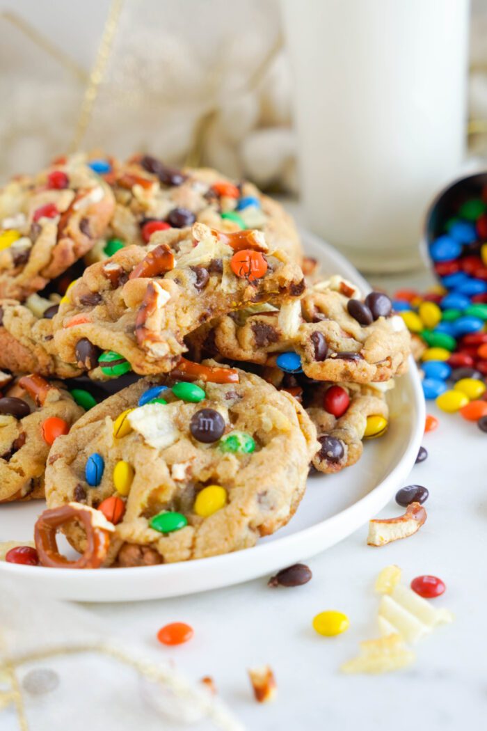 A plate of cookies filled with colorful candy pieces and pretzels, surrounded by scattered candy, with a glass of milk in the background.