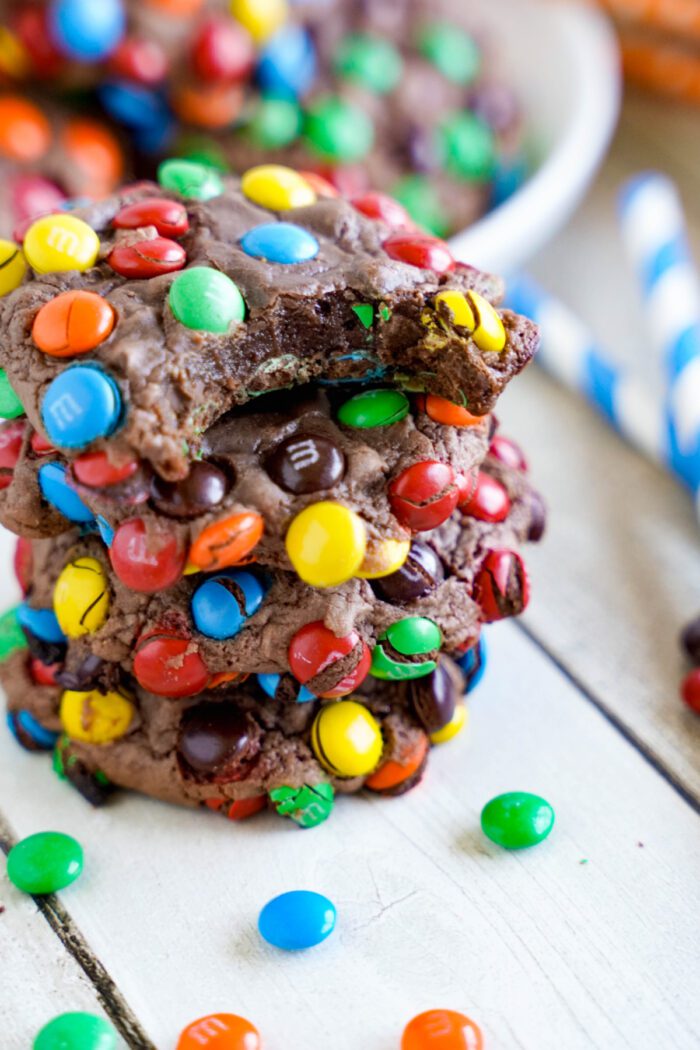 A stack of four chocolate cookies covered in colorful candy-coated chocolates, with one cookie on top showing a bite taken out of it. Scattered candies surround the cookies on a white wooden surface.