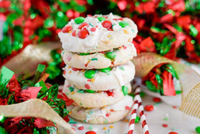 A stack of five Christmas cookies with white chocolate and colorful M&Ms, surrounded by red and green decorative ribbons and ornaments.