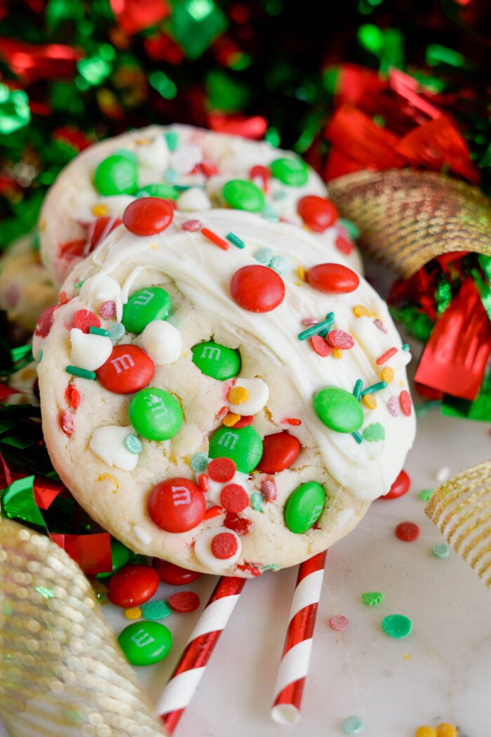 Two holiday-themed cookies topped with white chocolate, red and green M&M's, and colorful sprinkles, with festive red and green ribbon decorations in the background.