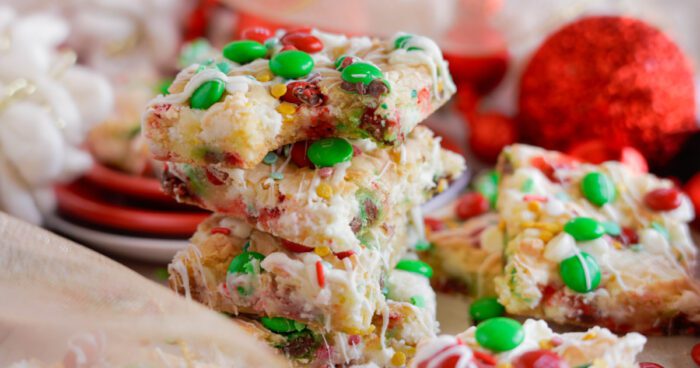 A stack of colorful Christmas Cookie bars with white frosting and multicolored candies, surrounded by festive decorations.