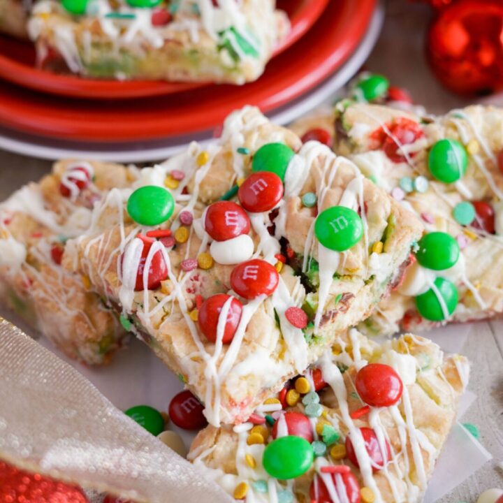 Festive cookie bars topped with red and green M&M's, white icing, and sprinkles are arranged on a plate and a table.