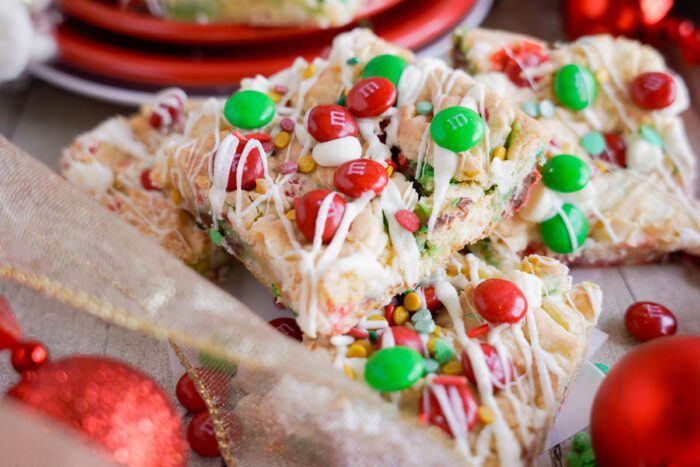 Christmas cookie bars topped with red and green M&Ms, white drizzle, and colorful sprinkles, placed on a table with red ornaments and a festive ribbon.