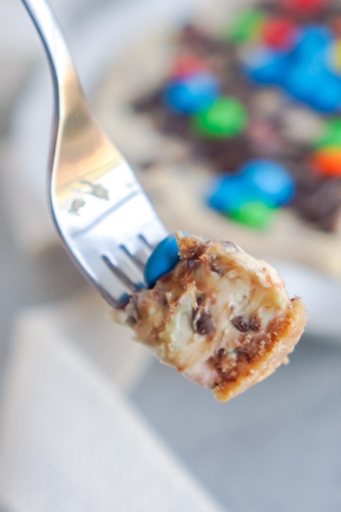 A fork holds up a bite of a dessert with layers of creamy filling, nuts, and chocolate pieces, with a colorful topping visible in the background.