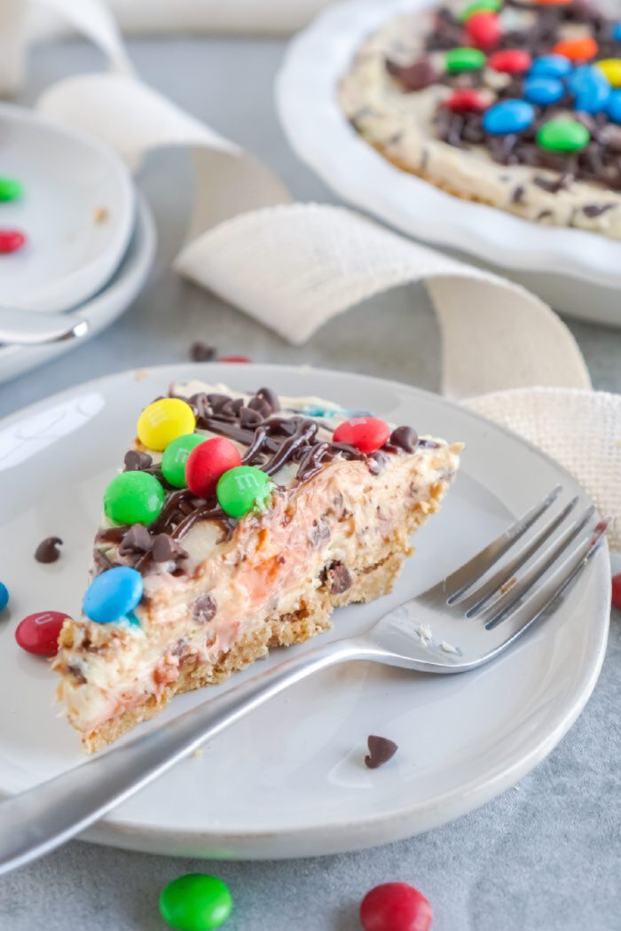 A slice of colorful M&M cheesecake topped with chocolate chips on a white plate with a fork beside it. The whole cheesecake is in the background.