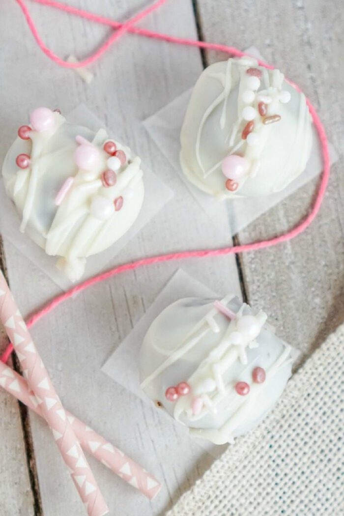 Three white chocolate-covered Oreo balls decorated with pink and white pearls on a wooden surface with pink ribbons.