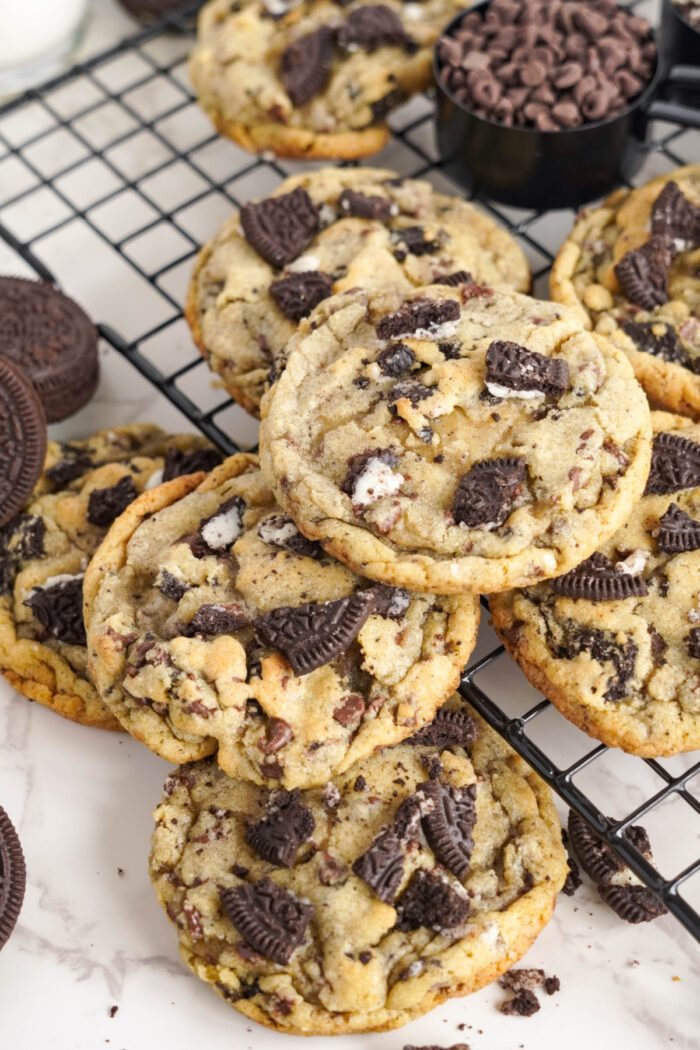 Freshly baked chocolate chip cookies with chunks of oreo cookies on a cooling rack, surrounded by ingredients.