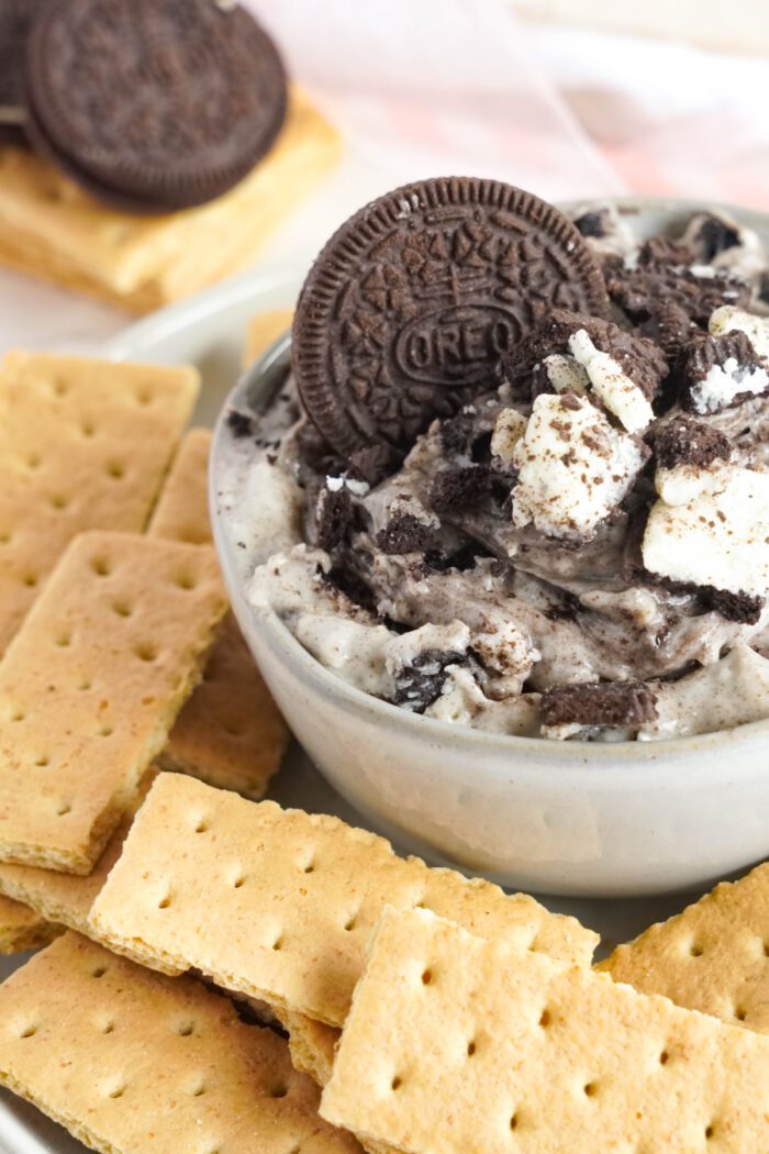 Bowl of Oreo dip topped with crushed Oreo cookies, surrounded by whole and crushed graham crackers.