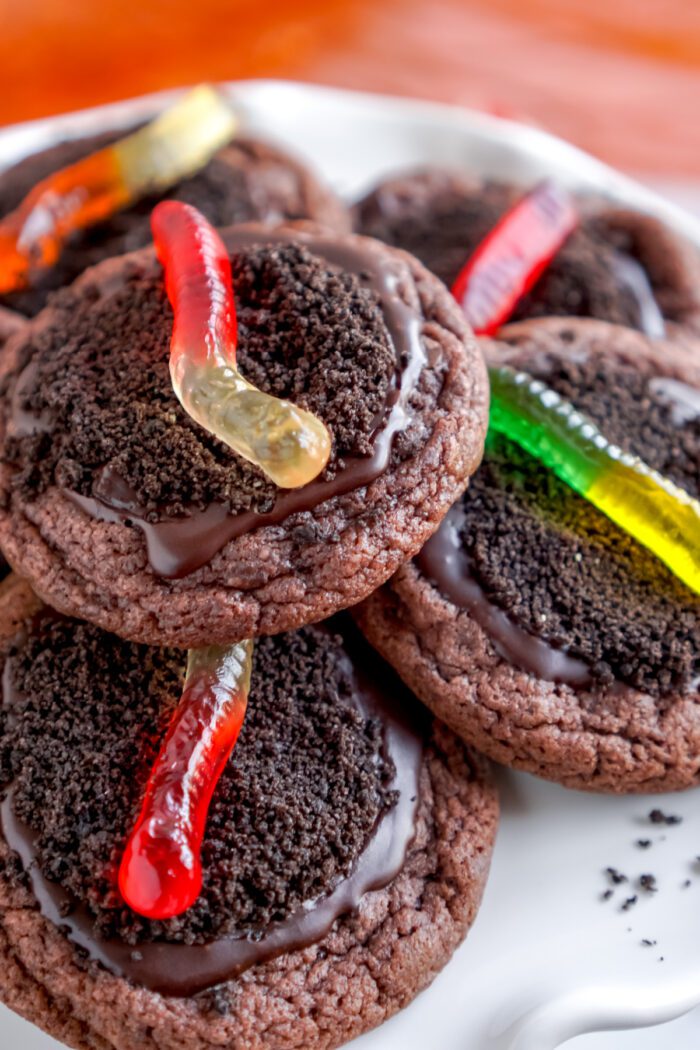 A stack of chocolate cookies topped with chocolate frosting and gummy worms on top, served on a white plate.