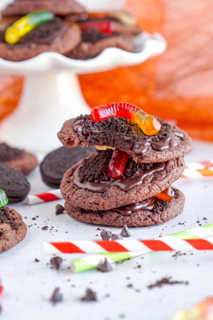 Stack of chocolate cookies with chocolate frosting and topped with gummy worms, surrounded by scattered candies and cookies on a white background.