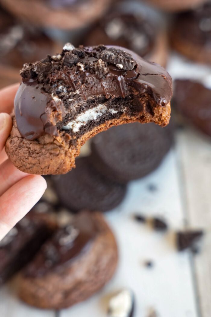 A hand holding a chocolate cookie with a bitten section revealing an Oreo center, topped in melted chocolate, with more cookies and crumbs in the background.