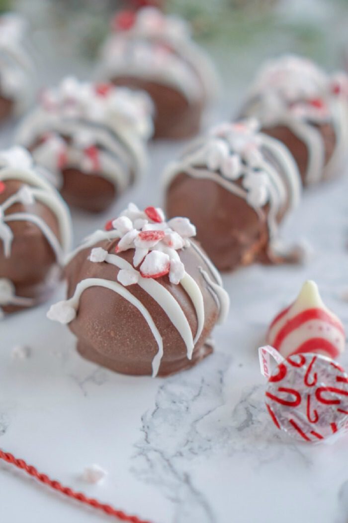 Close-up of several round chocolate peanut butter balls topped with white icing and crushed peppermint pieces, arranged on a marble surface. A red and white Hershey's Kiss is partially visible next to them.