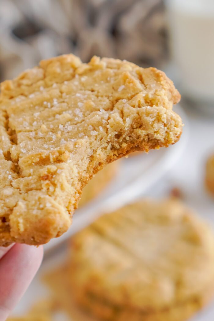 A close-up of a bitten peanut butter cookie held in someone's hand. The cookie is coated with sugar crystals.