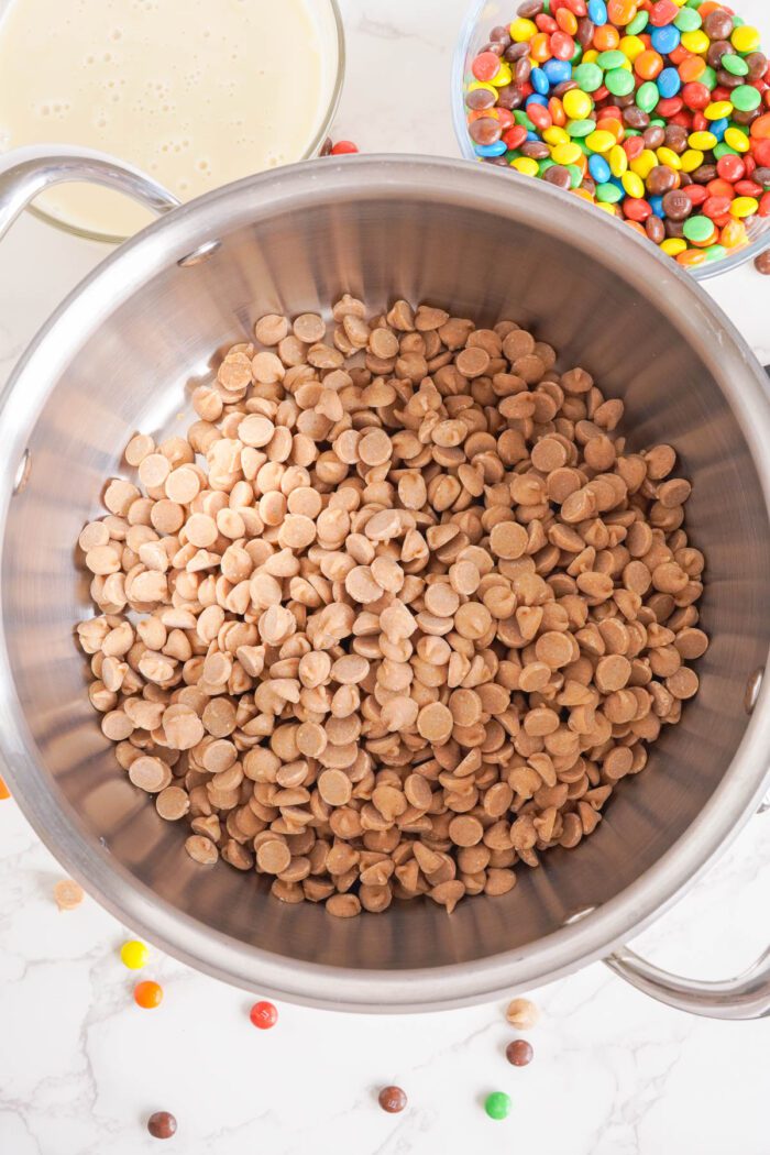 A stainless steel bowl filled with peanut butter baking chips, next to a bowl of sweetened condensed milk and a bowl of mini M&M candies, on a marble countertop.