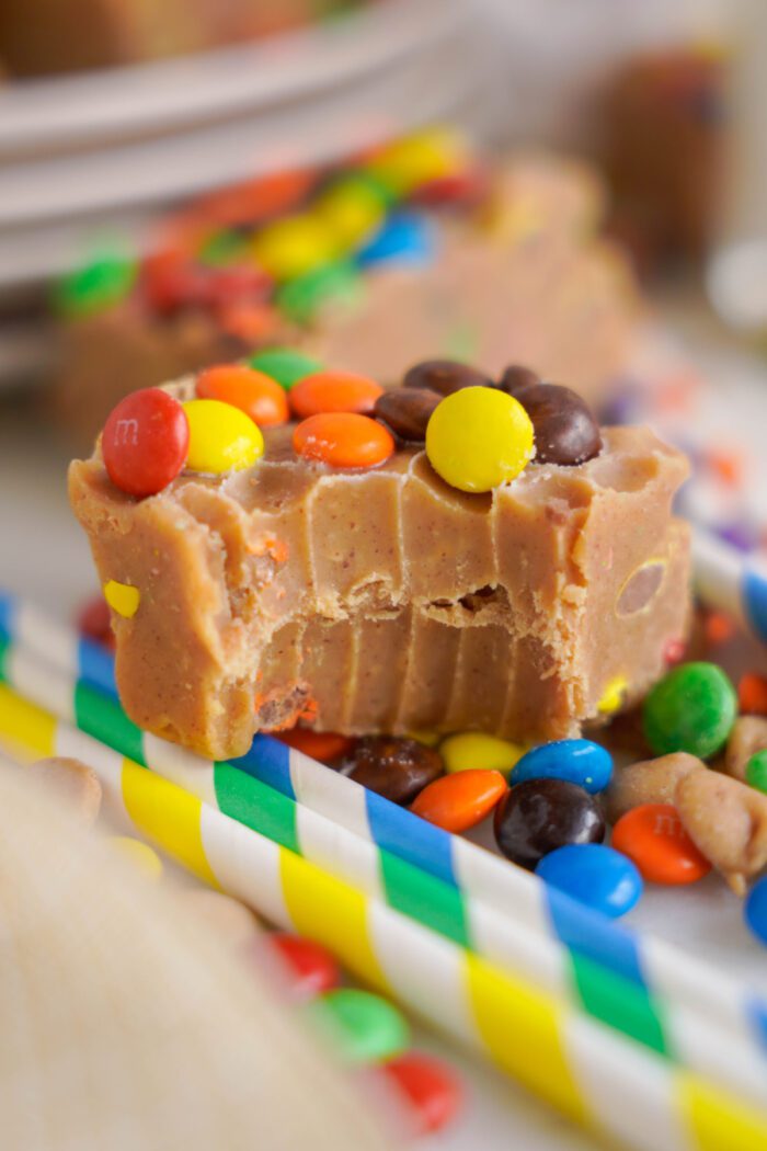 Close-up of a piece of peanut butter fudge with colorful candy-coated chocolates on top, partially eaten, placed on a striped napkin.