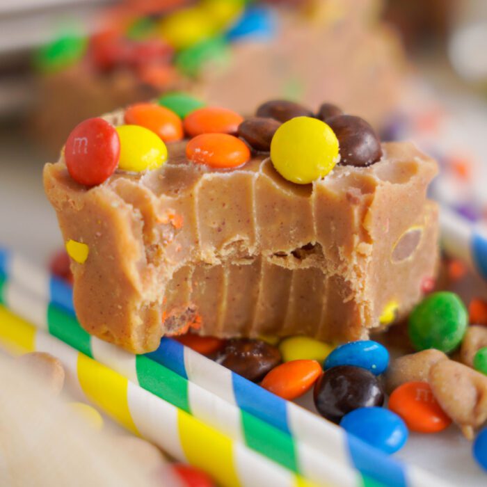 A close-up image of a slice of peanut butter fudge topped with colorful m&m candies, resting on a bed of more m&ms.