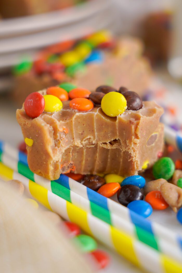 A piece of fudge with colorful candy-coated chocolates on top, showcasing a bite taken out of it, rests on a plate with other similar pieces in the background.