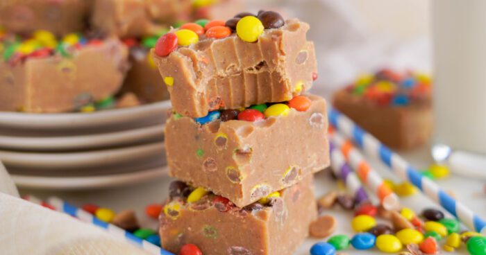 Stacked peanut butter fudge pieces with M&Ms on a plate, surrounded by extra M&Ms on a white surface.