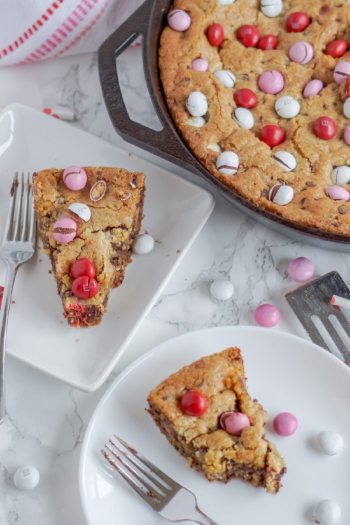 Two slices of M&M skillet cookie topped with red, pink, and white candies are placed on white plates with forks. The remaining skillet cookie is in a pan, with candies visible on the surface.