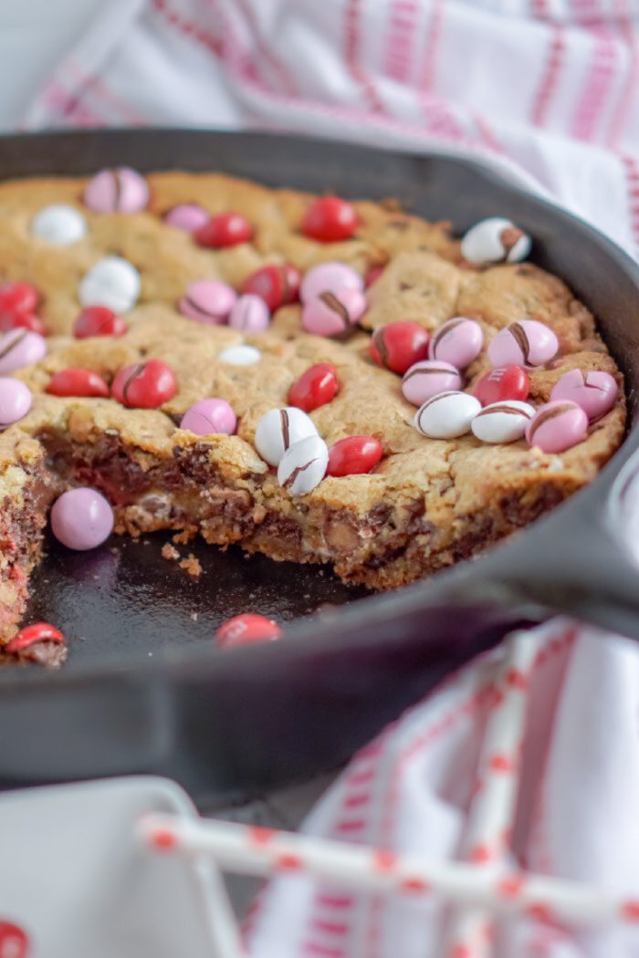 A skillet M&M cookie with a slice missing, topped with red, pink, and white candy pieces, sits on a white towel with red stripes.
