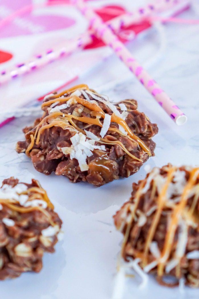 Close-up of no-bake chocolate coconut cookie drizzled with caramel on a marble surface, with pink striped straws and a pink heart-patterned cloth in the background.