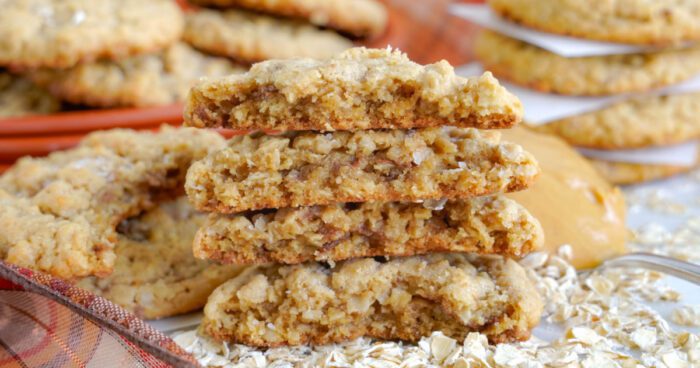 A close-up of several stacked oatmeal cookies with crispy edges and chewy centers, surrounded by oats and more cookies in the background.