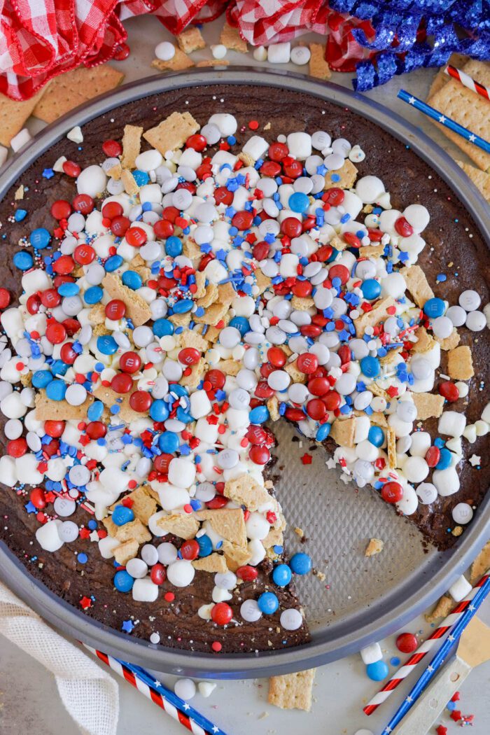 A round dessert brownie pizza on a pan, covered with white frosting, and topped with red, white, and blue candies and marshmallows. A slice is missing from the dessert.