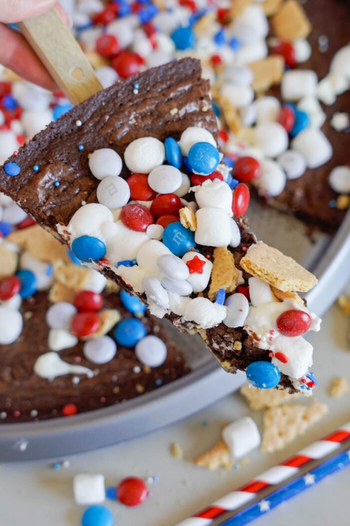 A slice of dessert pizza topped with white, red, and blue candies, mini marshmallows, graham cracker pieces, and icing, being lifted from a larger section.