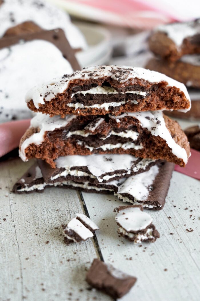 A stack of chocolate cookies stuffed with Oreos on a wooden table, surrounded by cookie crumbs and pieces.
