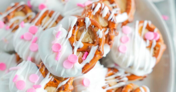 Pretzels partially dipped in white chocolate and decorated with pink sprinkles are arranged in a pile.