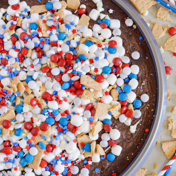 A tray of brownie pizza dessert topped with red, white, and blue candies, miniature marshmallows, graham cracker pieces, and sprinkles on a brown base.