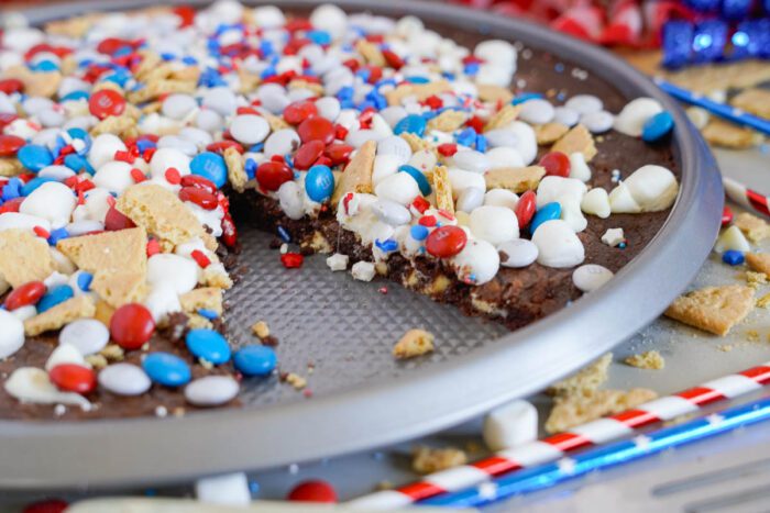 Round tray with a partially eaten dessert pizza topped with red, white, and blue candy-coated chocolates, white marshmallows, and pieces of graham cracker.