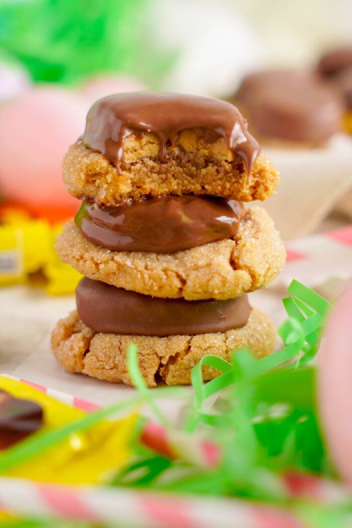 A stack of three peanut butter cookies, each coated with a layer of melted chocolate, sits on a table surrounded by colorful candy wrappers and decorative green grass.