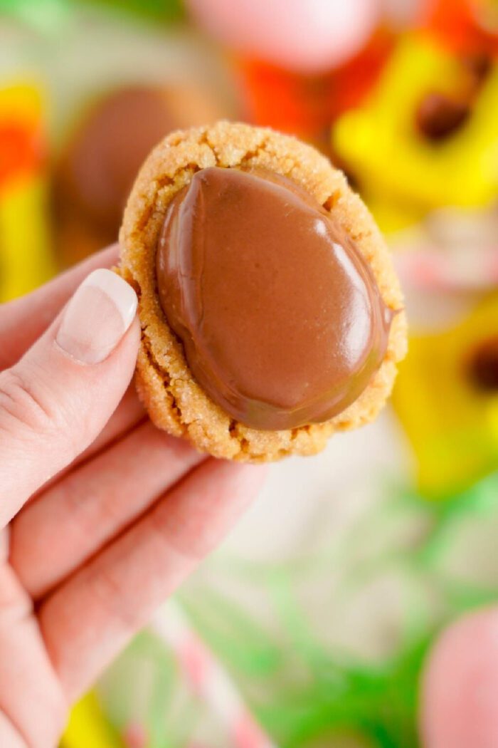 A hand holds a peanut butter cookie topped with a chocolate egg. Brightly colored flowers are blurred in the background.