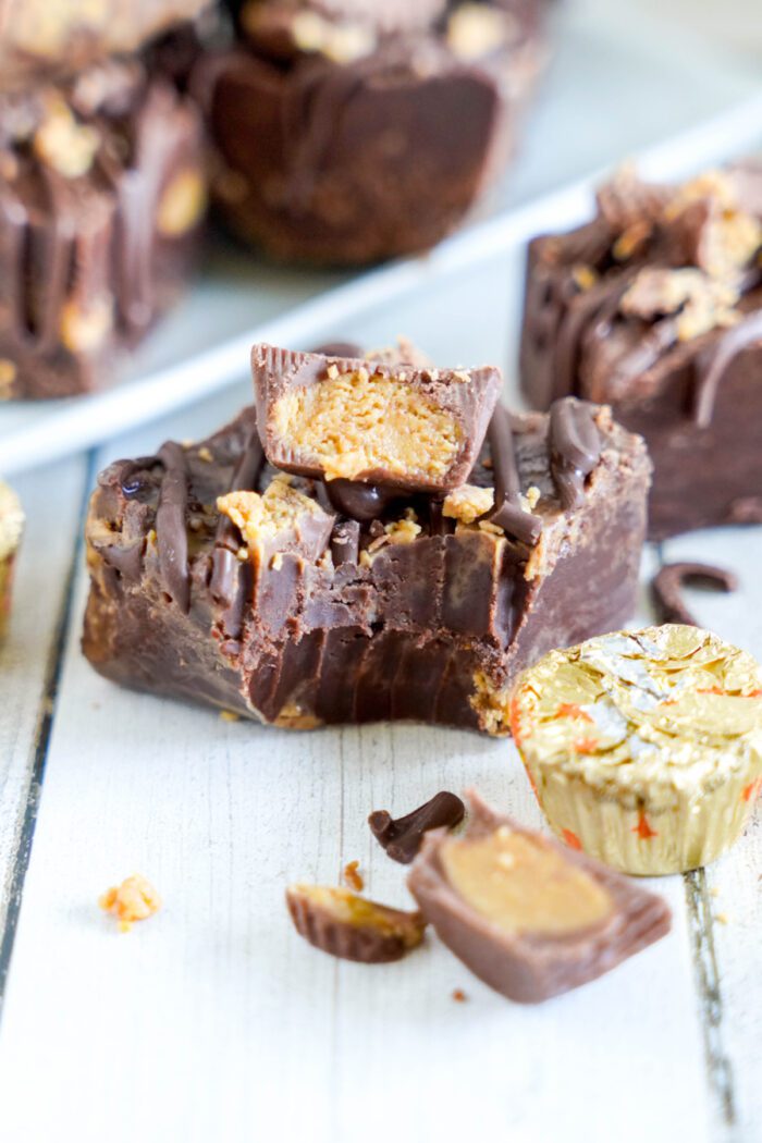 Close-up of chocolate squares with peanut butter filling, some stacked and partially eaten. A small, wrapped peanut butter cup is nearby on a white wooden surface.