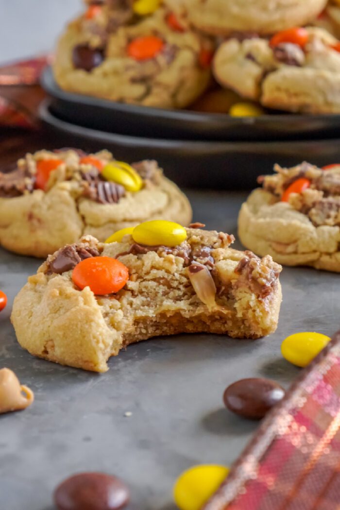 Close-up of peanut butter cookies topped with Reese's Pieces candies. One cookie in the foreground has a bite taken out of it. Background shows more cookies on a black plate.