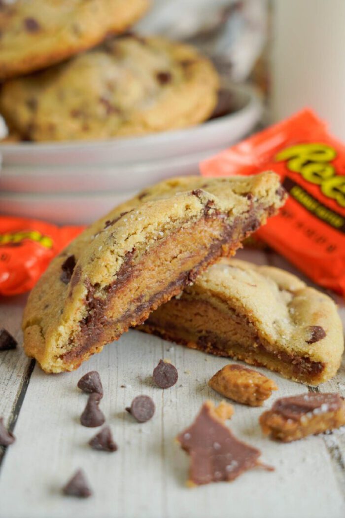 A chocolate chip cookie split open to reveal a Reese's peanut butter cup inside, with chocolate chips and more cookies in the background.