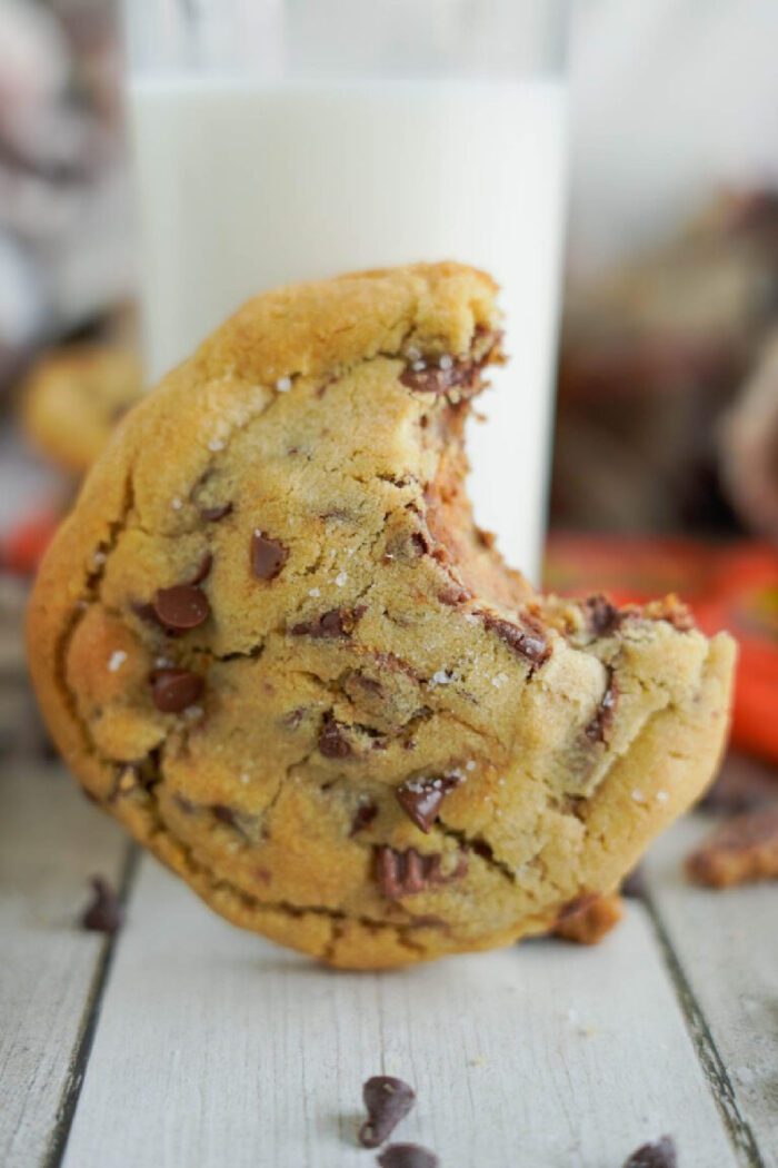 A reese's chocolate chip cookie with a bite taken out leans against a glass of milk on a wooden surface.