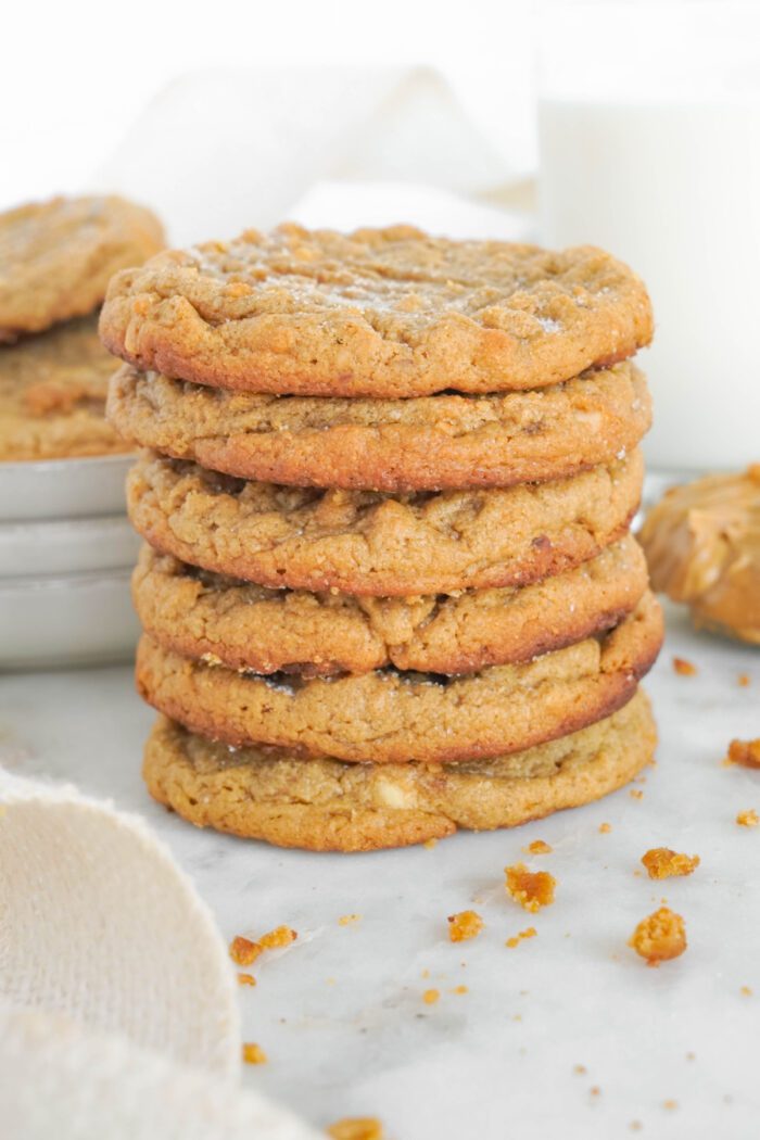 A stack of six peanut butter cookies is placed on a white surface with cookie crumbs scattered around. A glass of milk and more cookies are visible in the background.