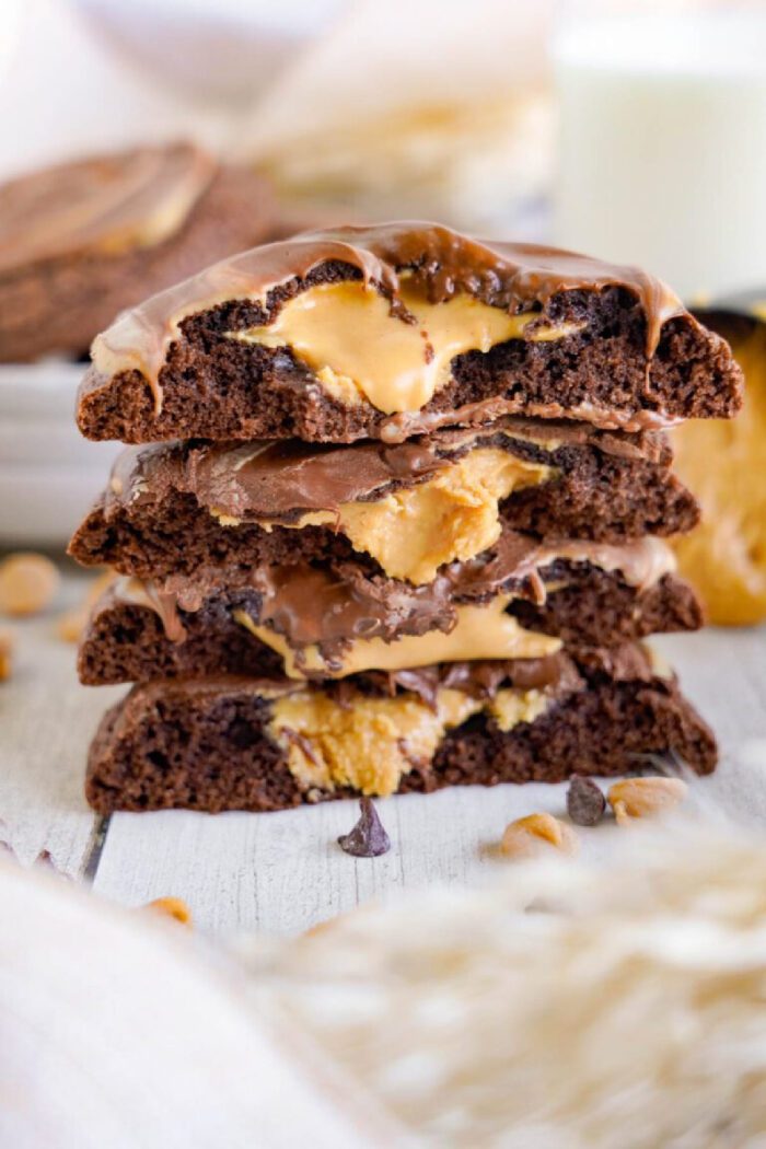 Four stacked chocolate cookies filled with a gooey peanut butter center, placed on a white wooden surface. A glass of milk and more cookies are blurred in the background.