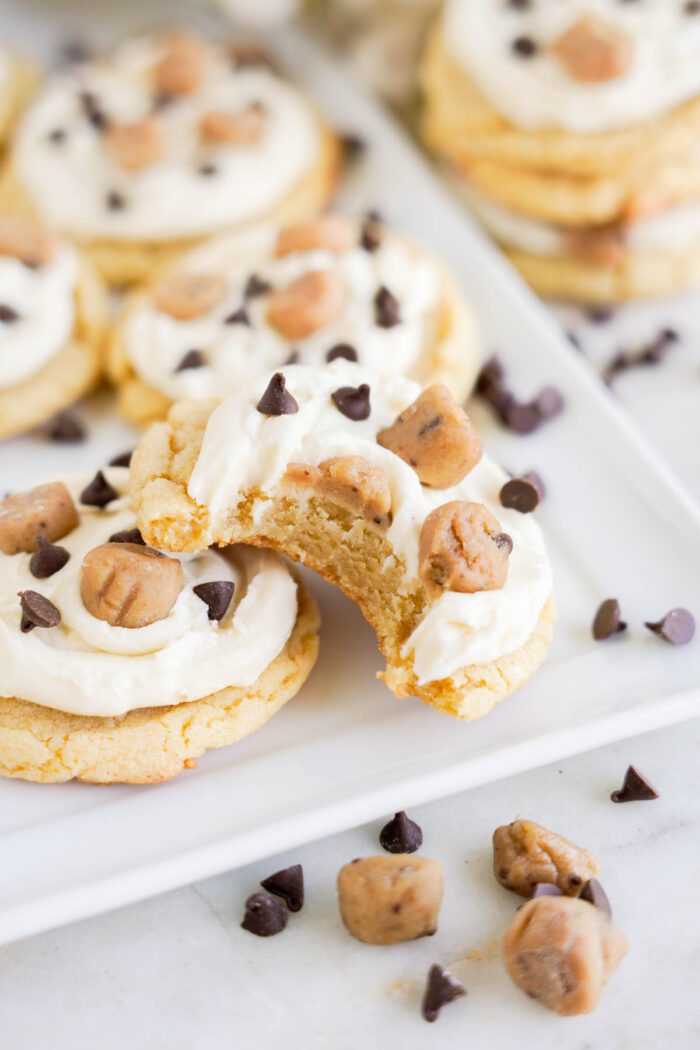 A close-up of cookies topped with frosting, chocolate chips, and small cookie dough pieces, displayed on a white rectangular plate. One cookie has a bite taken out of it, showcasing an easy cookie dough cookies recipe perfect for any occasion.